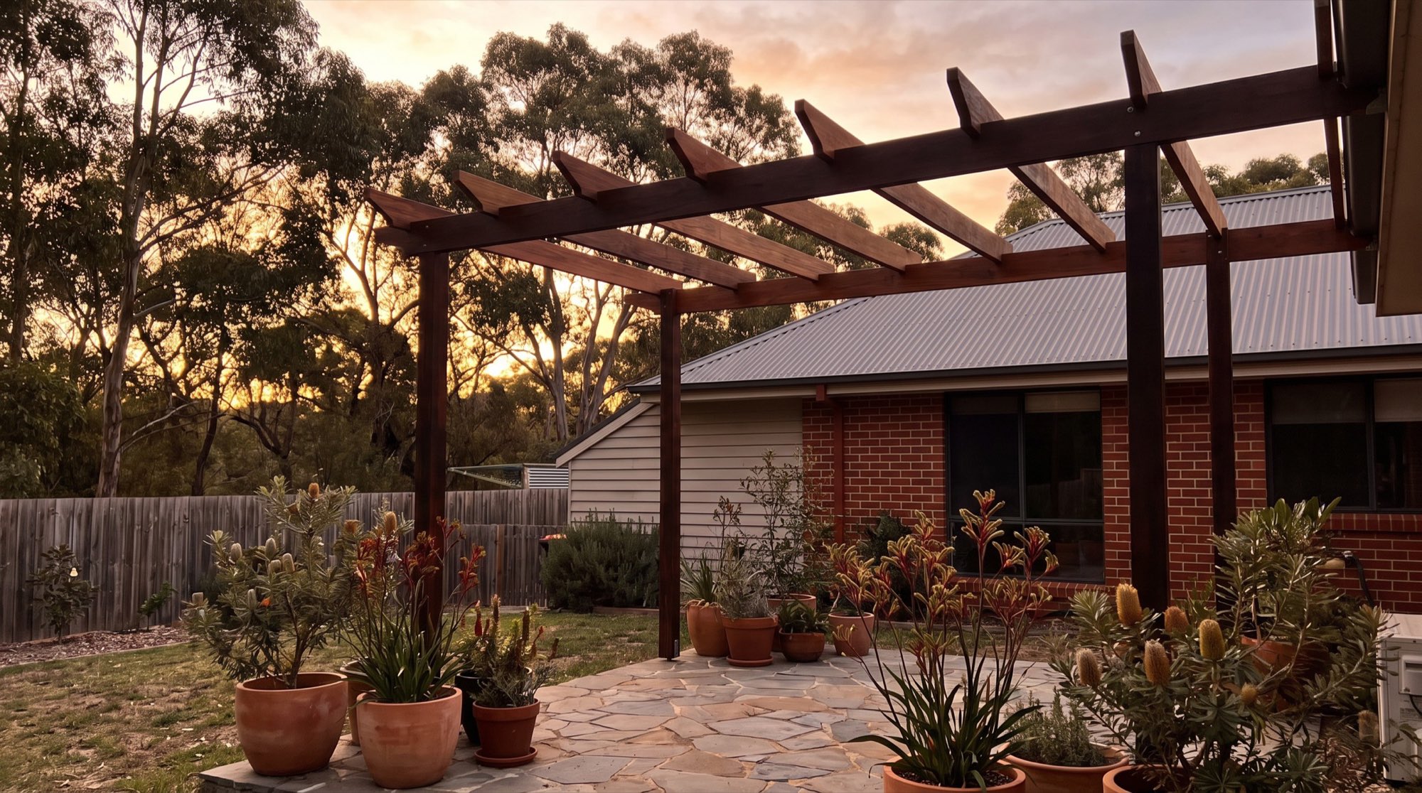 A timber pergola over a paved courtyard with potted plants beside a brick home