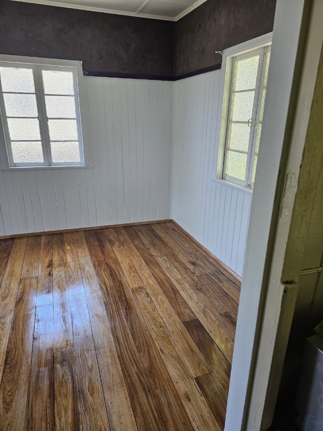 Polished hardwood floorboards meeting white timber-lined walls beside a window