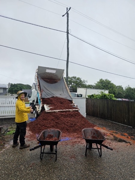 A person in a wet-weather jacket beside a tipper truck unloading garden mulch, with two wheelbarrows ready