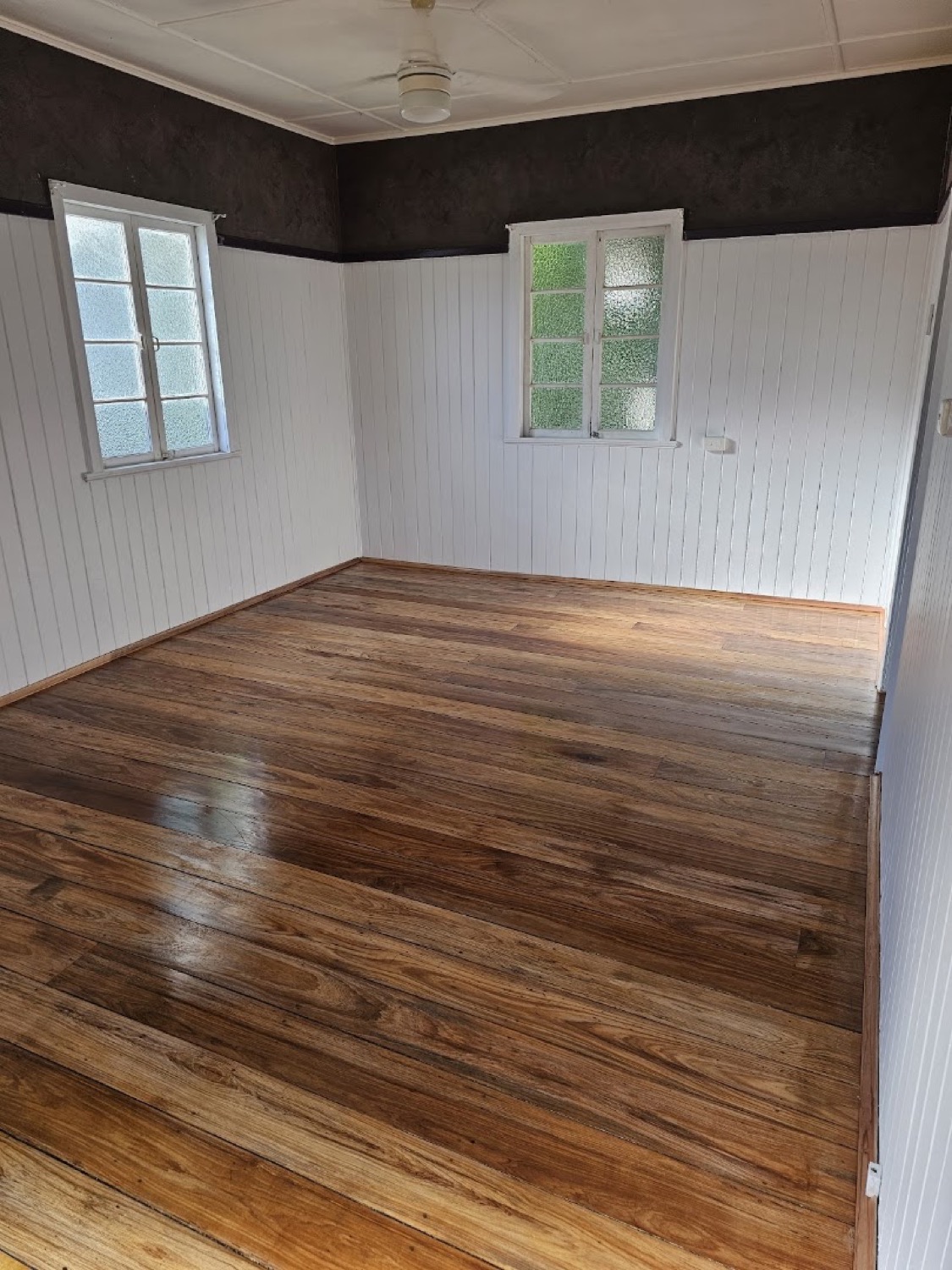 A room with polished timber floorboards, white timber-lined walls and two sash windows